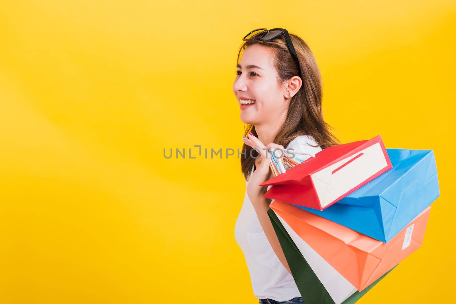 Asian Thai portrait happy beautiful cute young woman smiling stand with sunglasses excited holding shopping bags multi color looking side, studio shot isolated yellow background with copy space