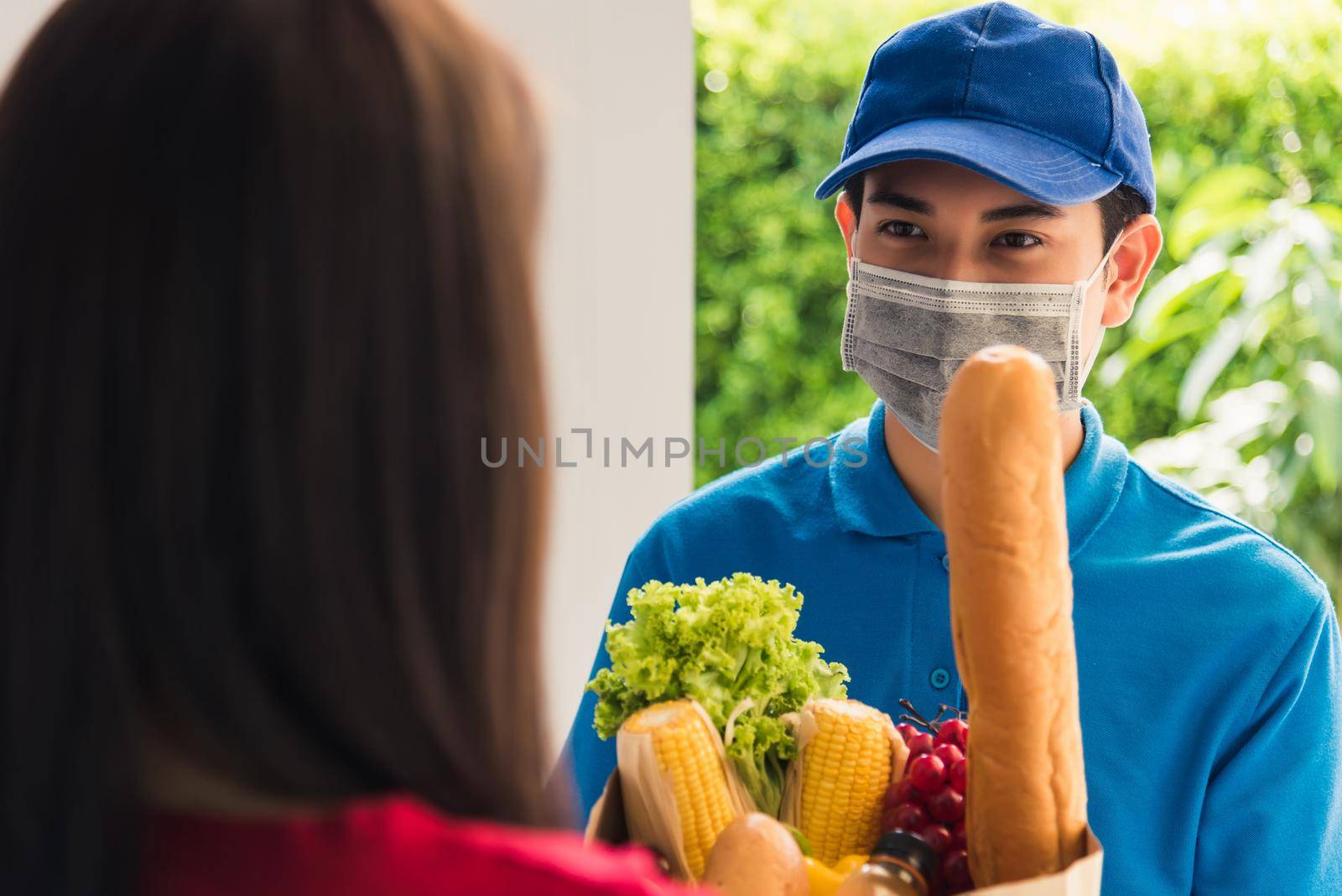 Asian young delivery man in uniform wear protective face mask he making grocery service giving fresh food to woman customer receiving front house under pandemic coronavirus, Back to new normal concept