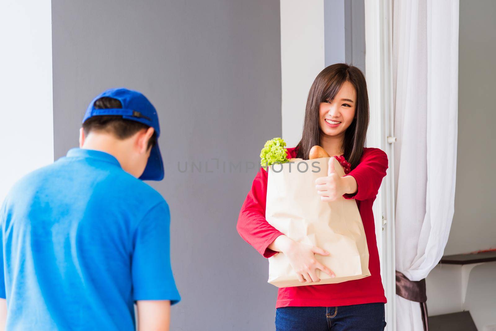 Asian young delivery man in blue uniform making grocery service giving fresh vegetables and fruits and food in paper bag to woman customer at front house after pandemic coronavirus, Back to new normal