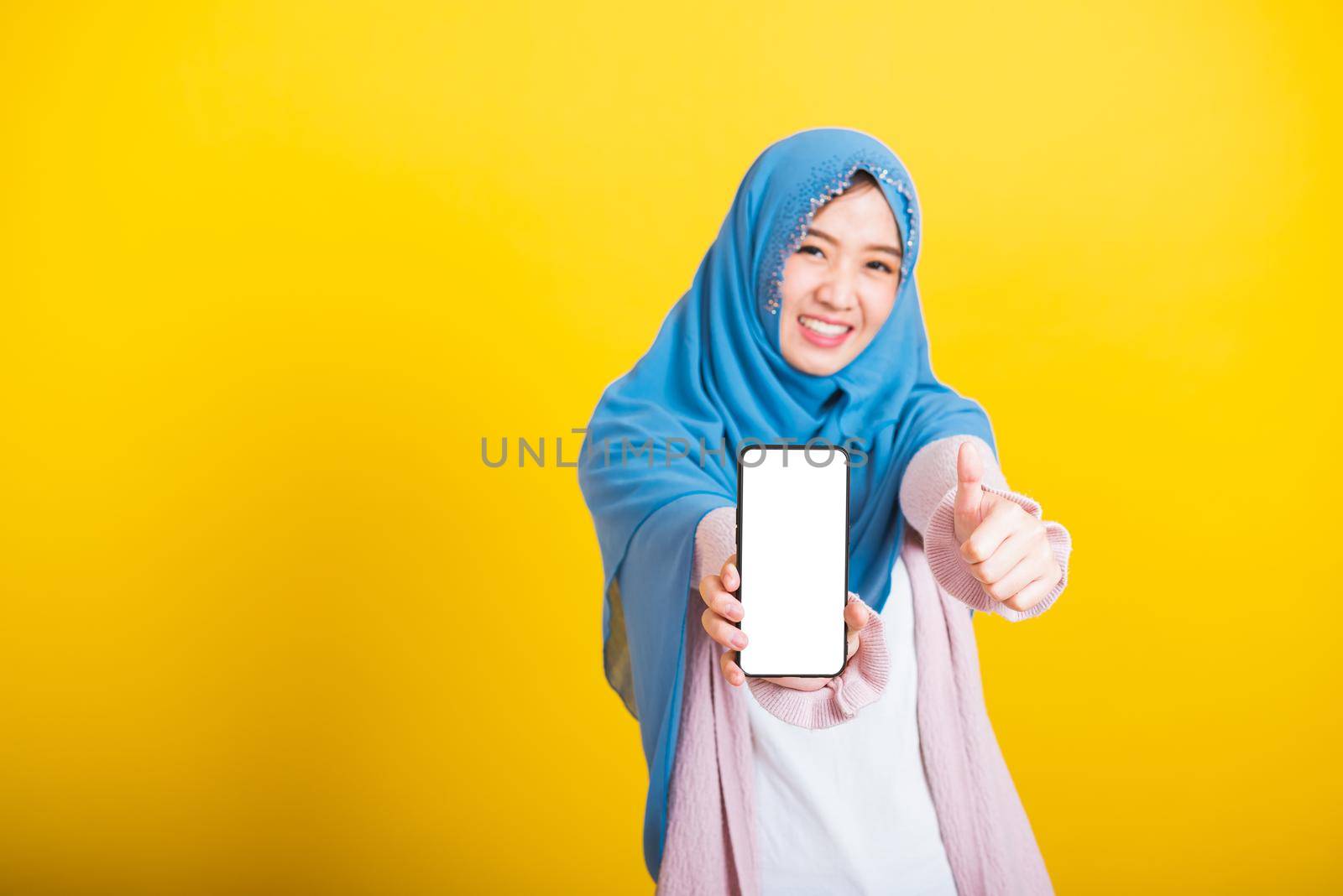 Asian Muslim Arab woman Islam wear veil hijab funny smile she showing blank screen smart mobile phone and point screen, studio shot isolated on yellow background