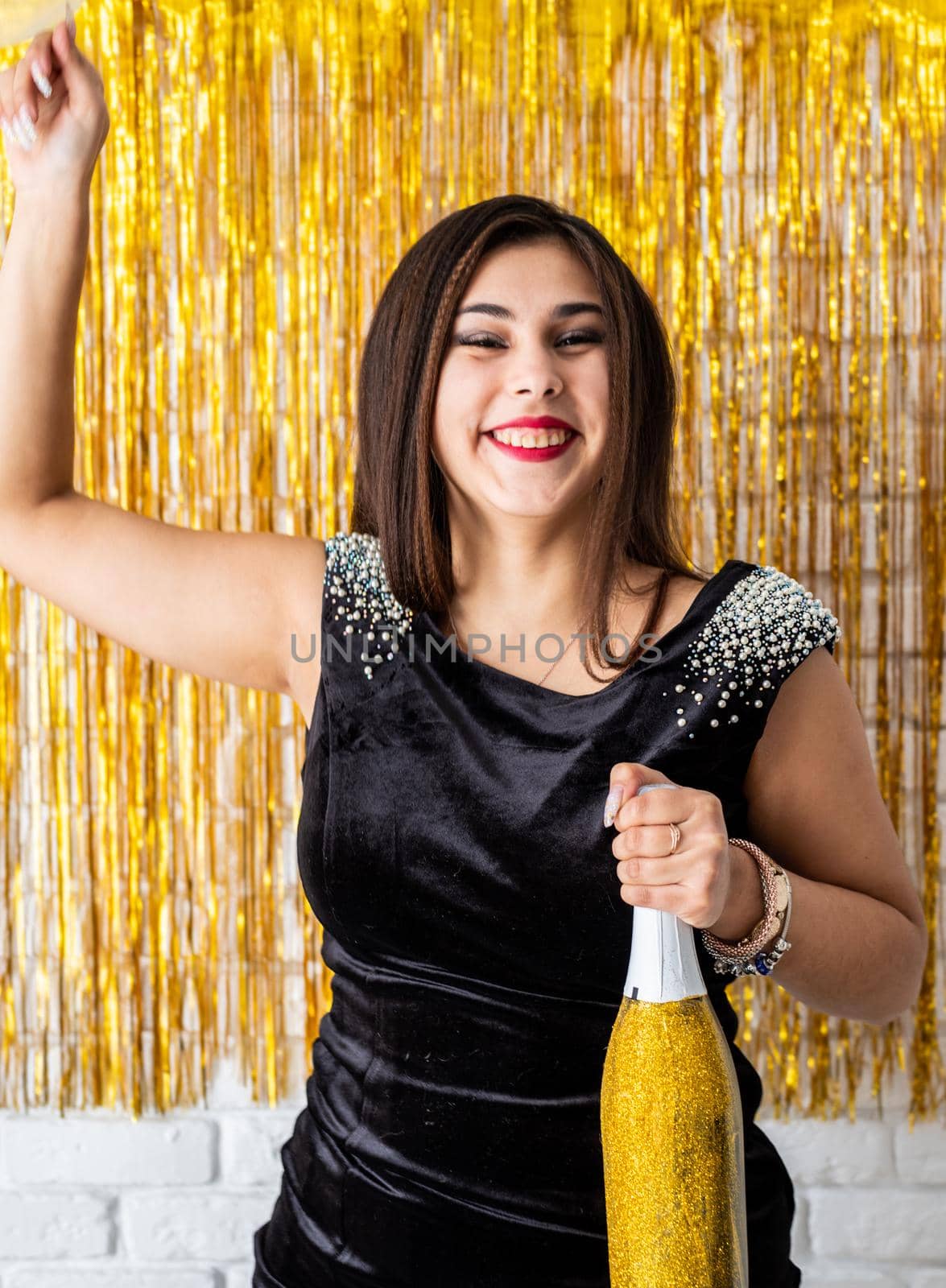 Beautiful brunette woman in black party dress celebrating her birthday holding balloon and champagne bottle by Desperada