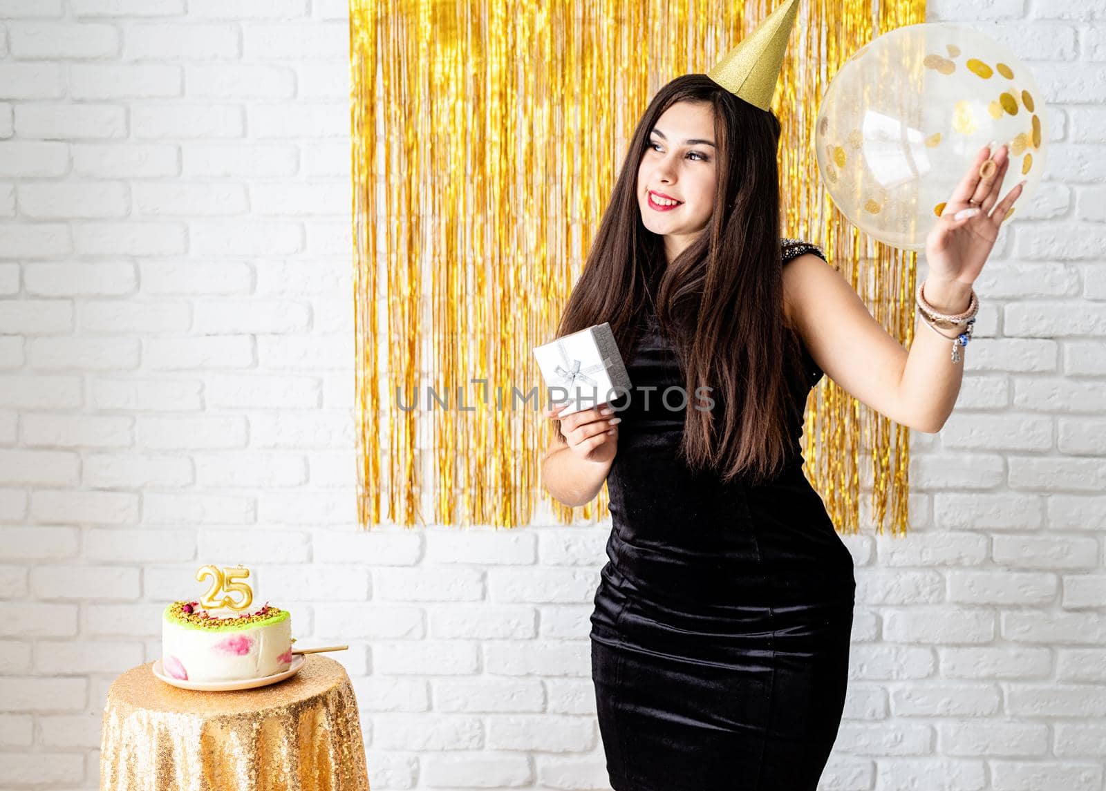 Birthday party. BBeautiful young woman in party dress and birthday hat holding balloon on golden background