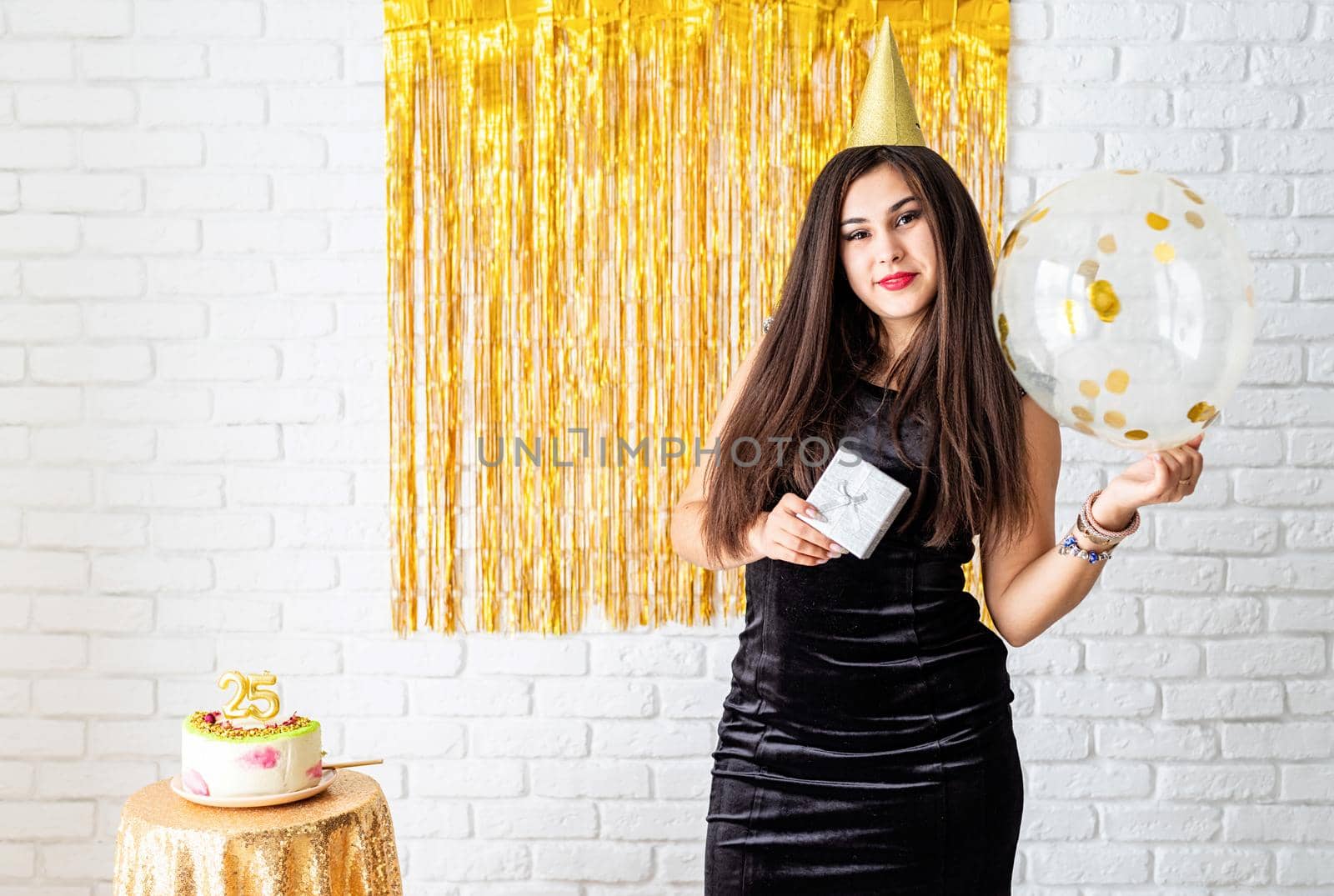 Birthday party. BBeautiful young woman in party dress and birthday hat holding balloon on golden background