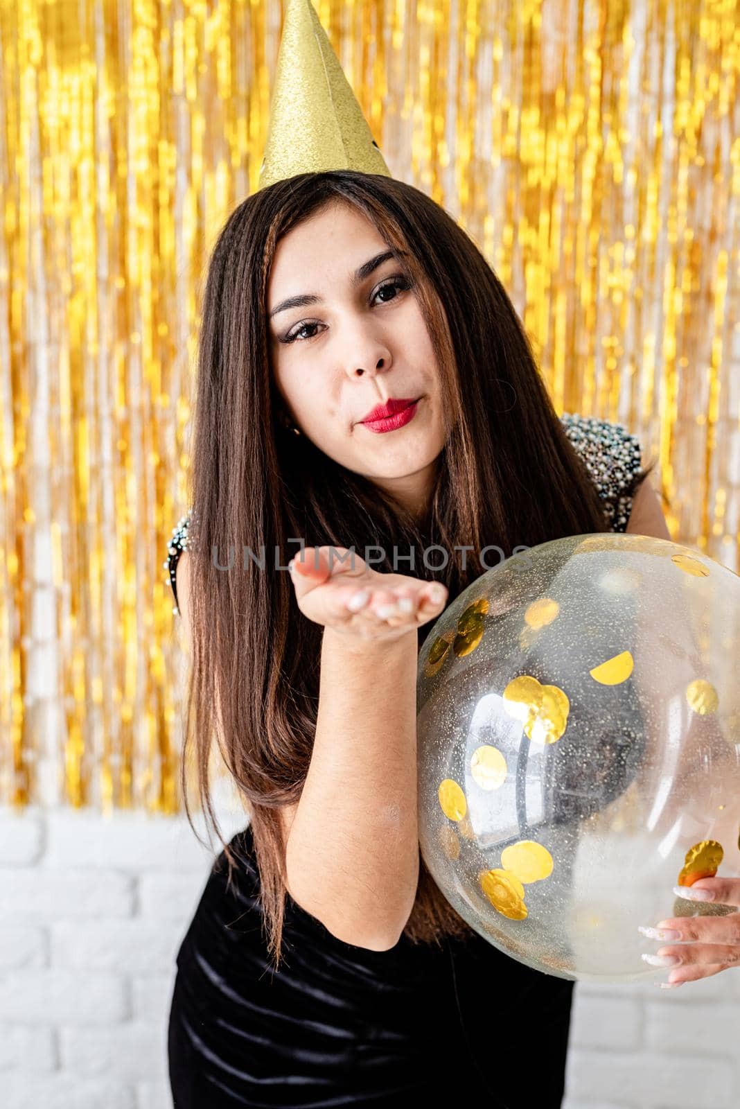 Birthday party. BBeautiful young woman in party dress and birthday hat holding balloon on golden background