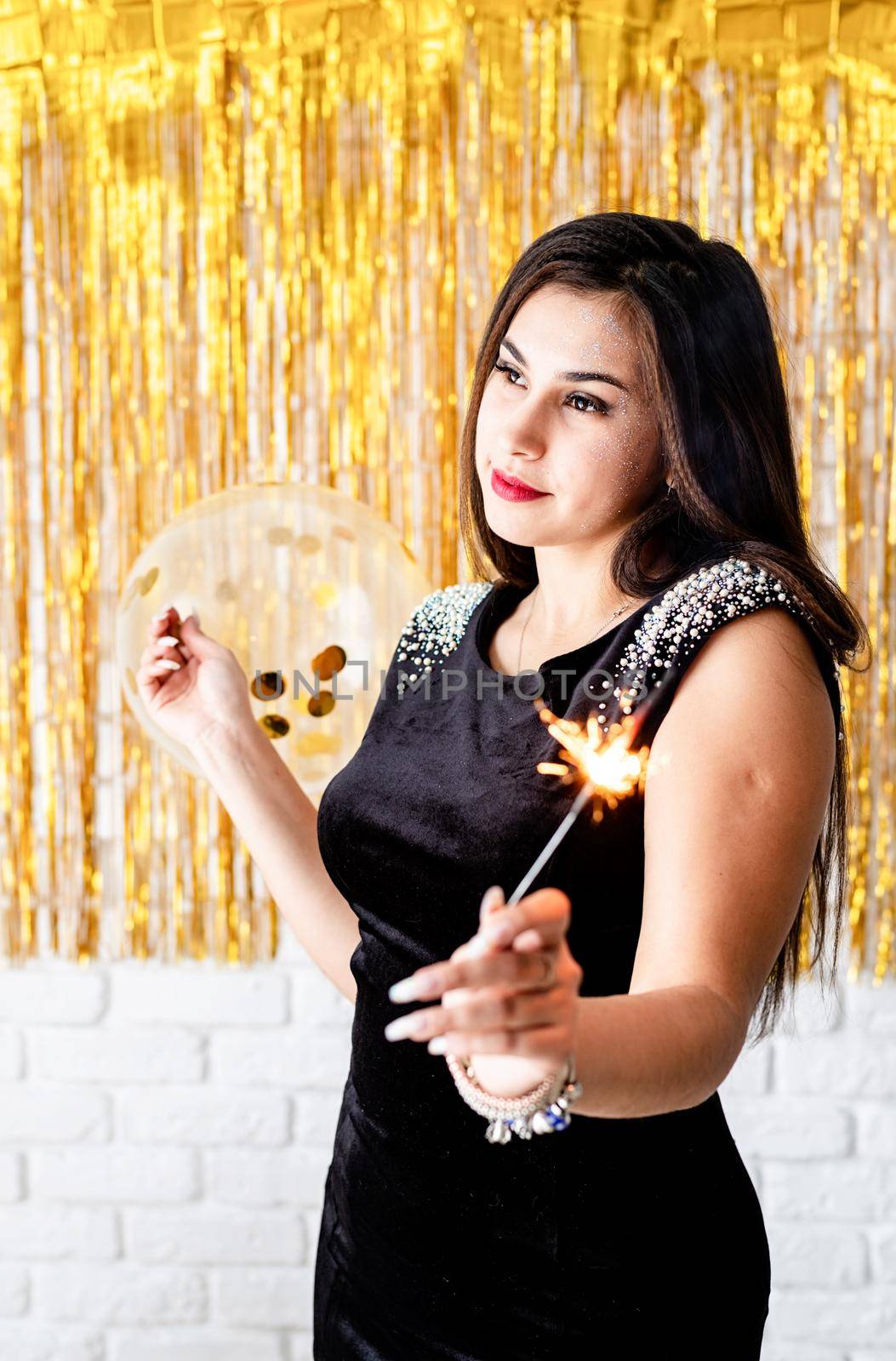 Birthday party. Beautiful young woman holding sparkler and balloon on golden background