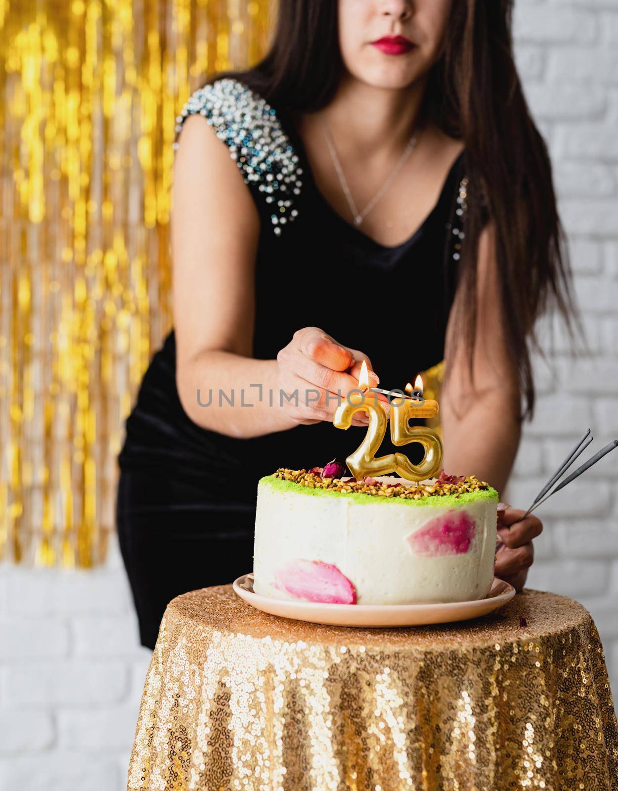 Beautiful brunette woman in black party dress celebrating her birthday lighting the candles on the cake by Desperada