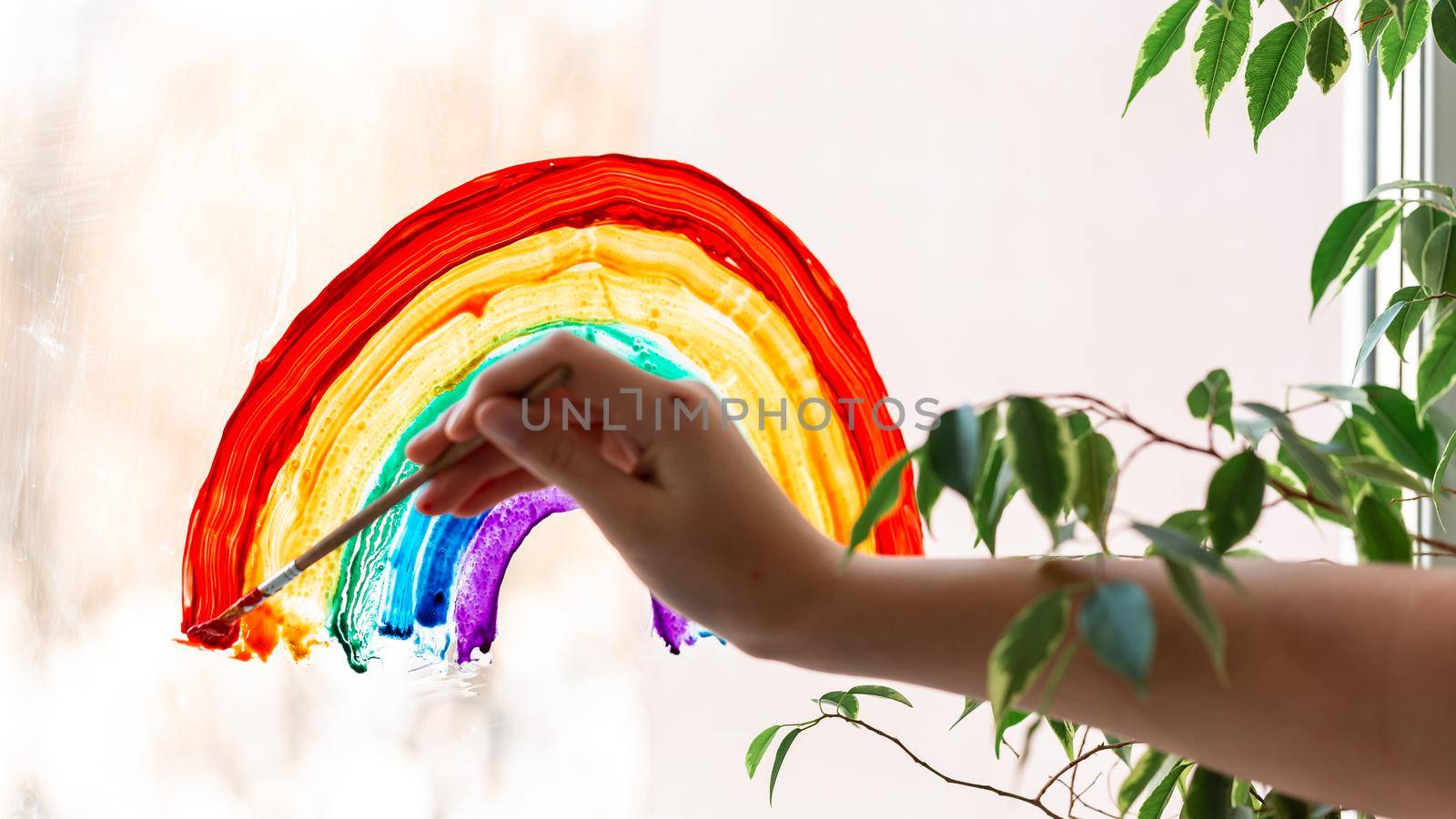 Little girl painting rainbow on window during Covid-19 quarantine at home. Stay at home social media campaign for coronavirus prevention, let's all be well, catch the rainbow