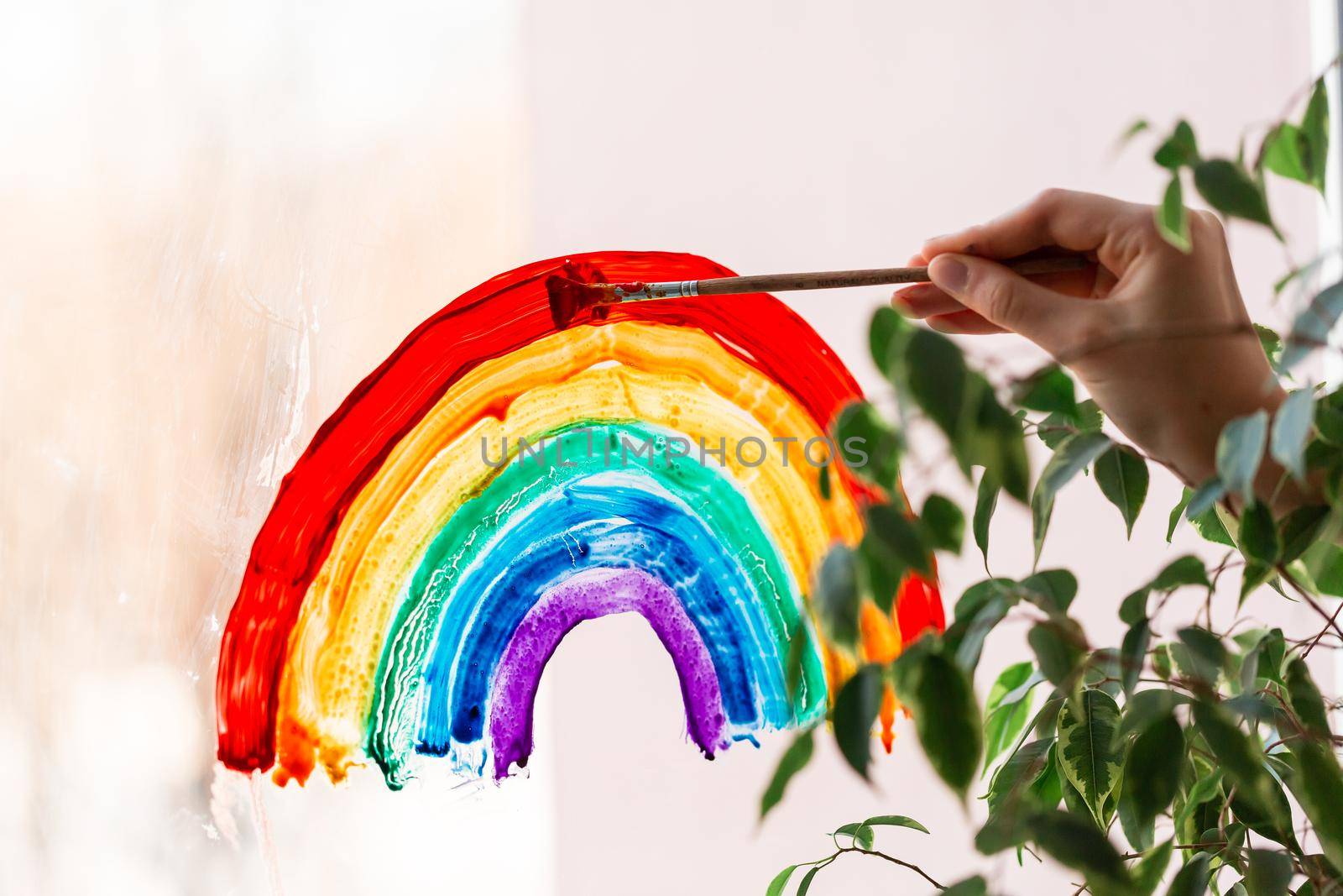 Little girl painting rainbow on window during Covid-19 quarantine at home. Stay at home social media campaign for coronavirus prevention, let's all be well, catch the rainbow