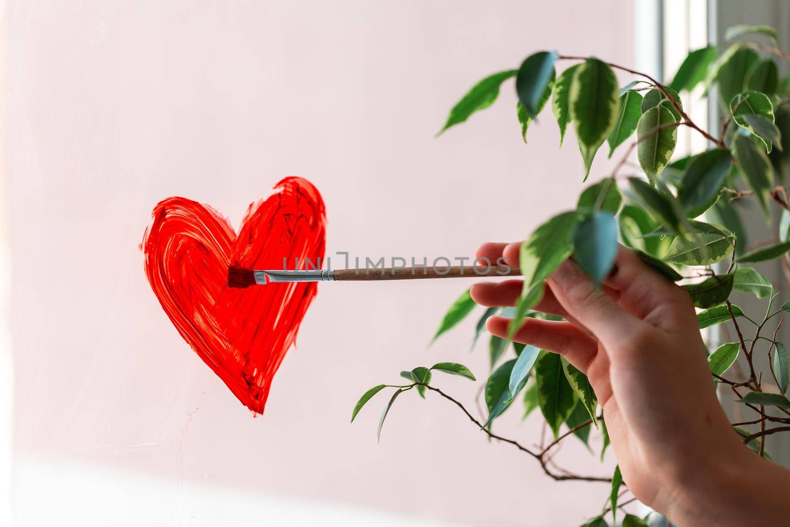 Little girl painting red heart on window during Covid-19 quarantine at home. Stay at home, stay healthy, let's all be well. Valentine's day concept.