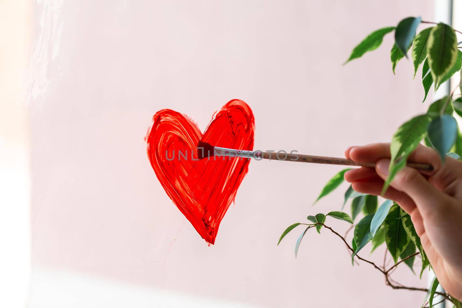 Little girl painting red heart on window during Covid-19 quarantine at home. Stay at home, stay healthy, let's all be well. Valentine's day concept.