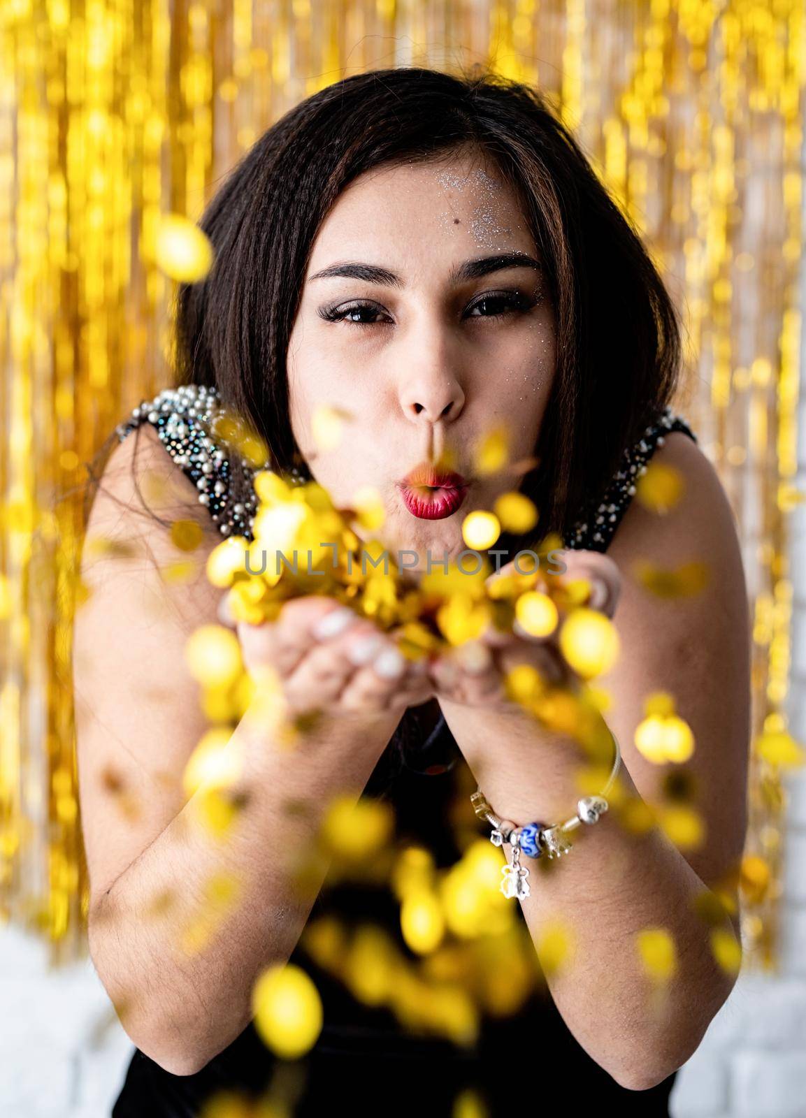 Birthday party. Portrait of young beautiful girl blowing golden confetti at holiday party