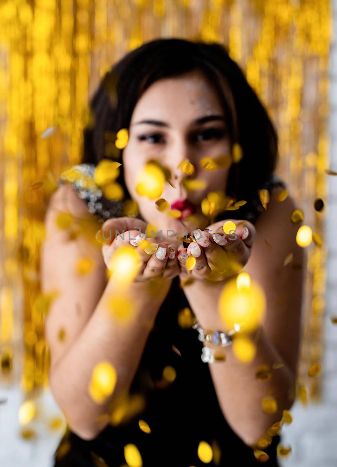 Portrait of young beautiful girl blowing golden confetti at holiday party by Desperada