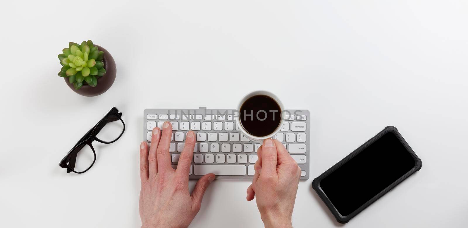 Office desk table with male hands holding cup of coffee over computer keyboard while working. Overhead with copy space available in flat lay format. 