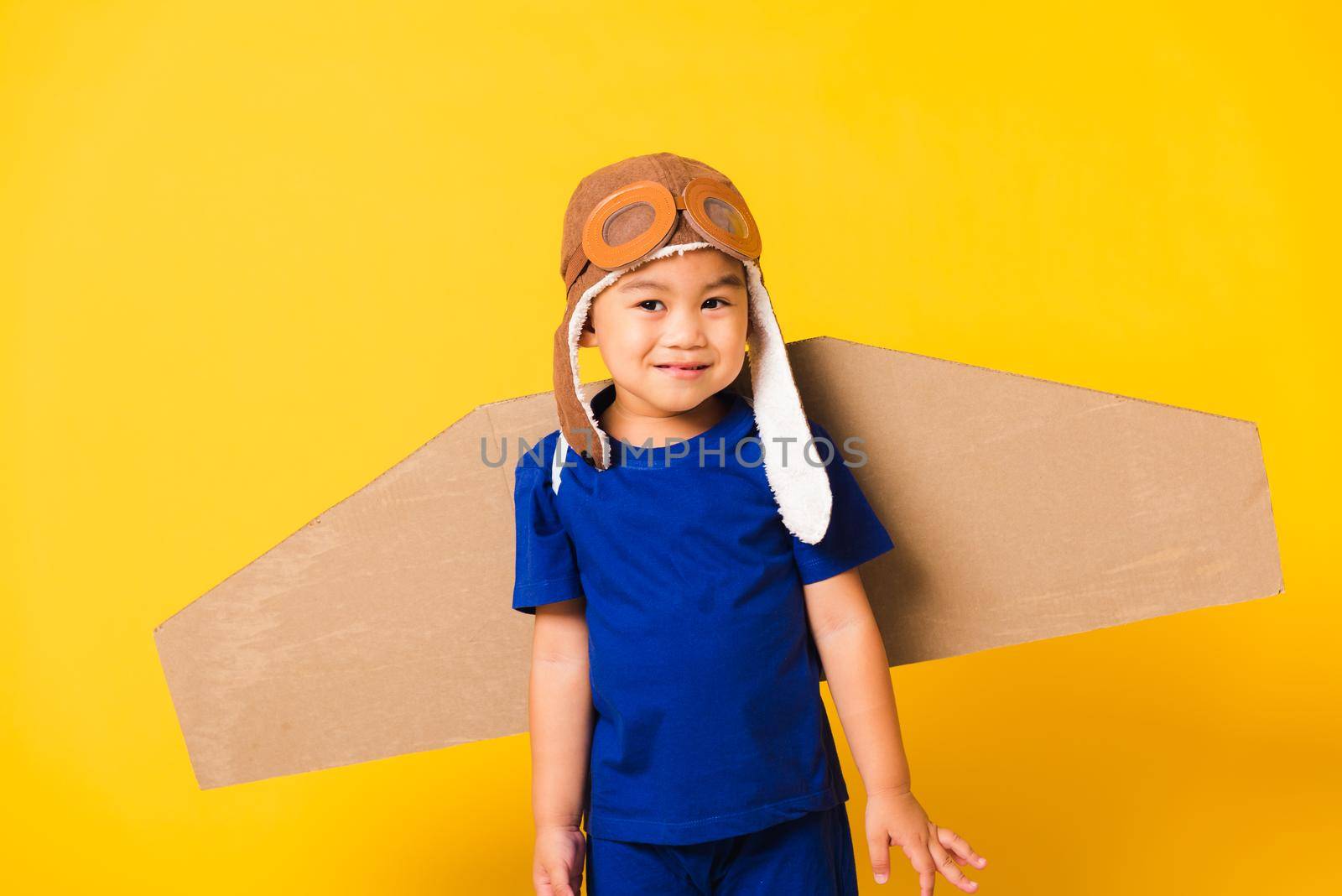 Happy Asian handsome funny child or kid little boy smile wear pilot hat playing and goggles with toy cardboard airplane wings flying, studio shot isolated yellow background, Startup freedom concept