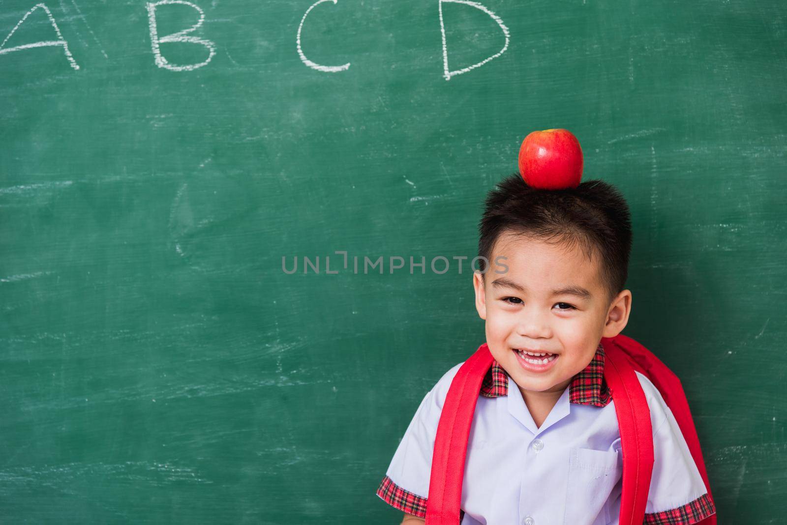 Child boy from kindergarten in student uniform with school bag and red apple on head smiling by Sorapop