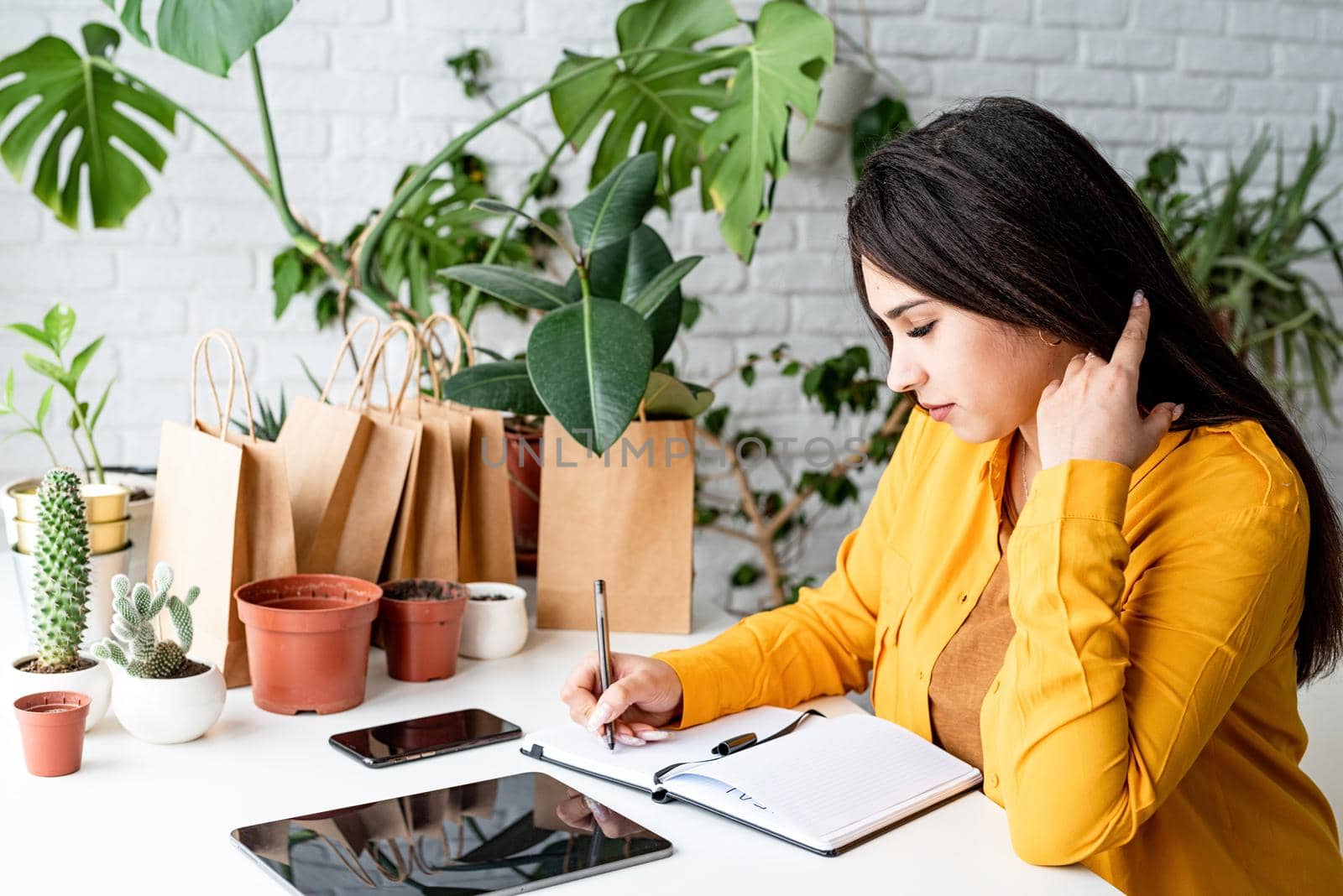 Home gardening. Small business. Young woman gardener making notes in the notepad surrounded with plants