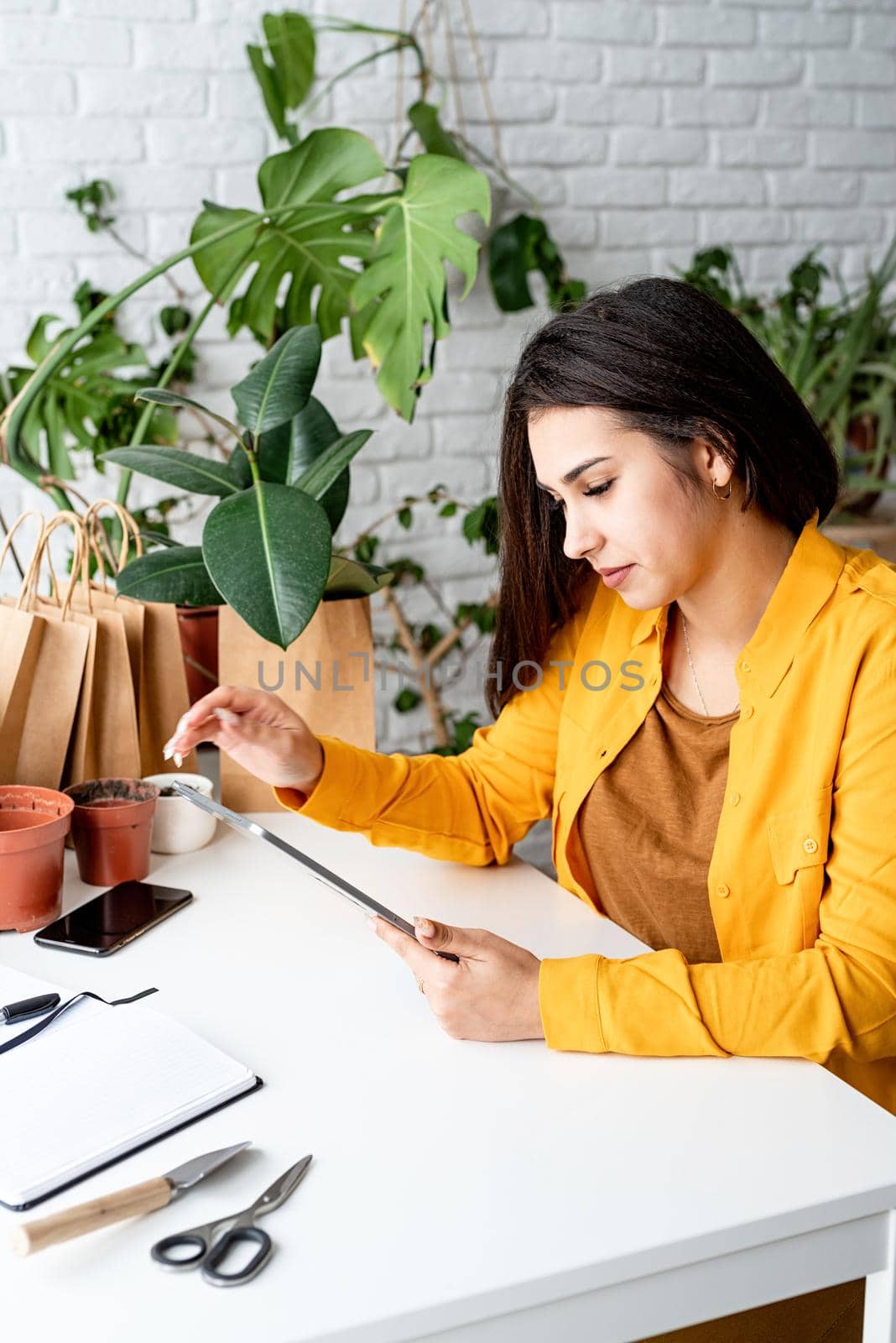 Home gardening. Small business. Woman gardener working on digital tablet surrounded with plants