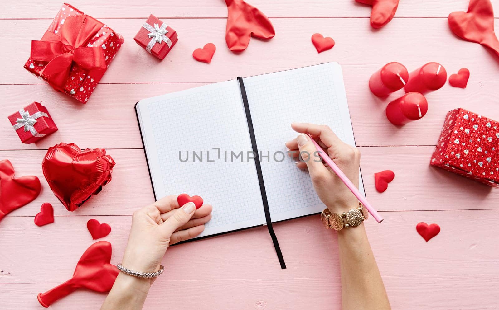 Valentines Day. Woman writing on blank paper sheets on pink wooden table decorated with red valentine candles and gifts