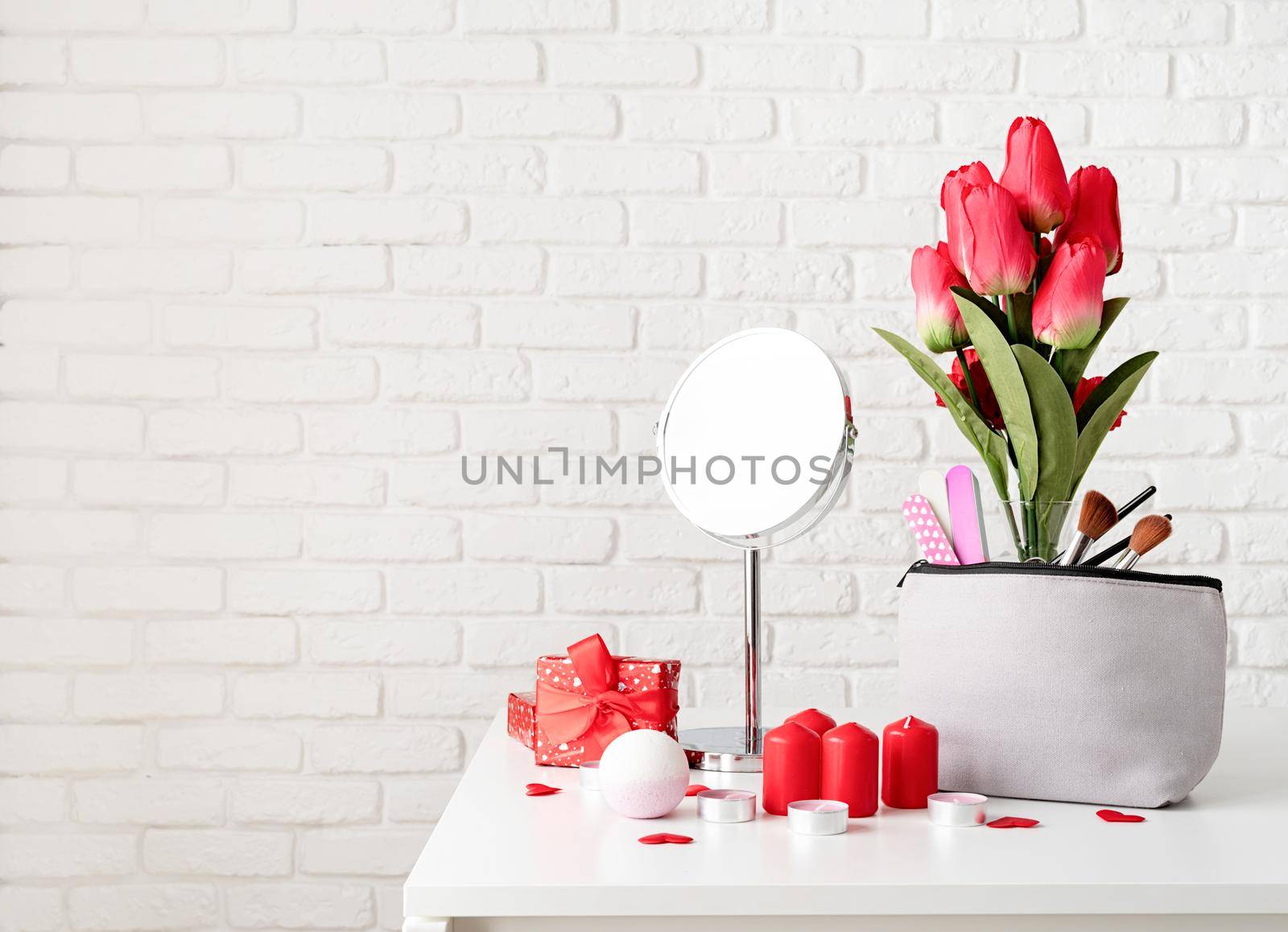 cosmetic and spa accessories with gift box, flowers and candles on dressing table front view on white brick wall background with copy space by Desperada