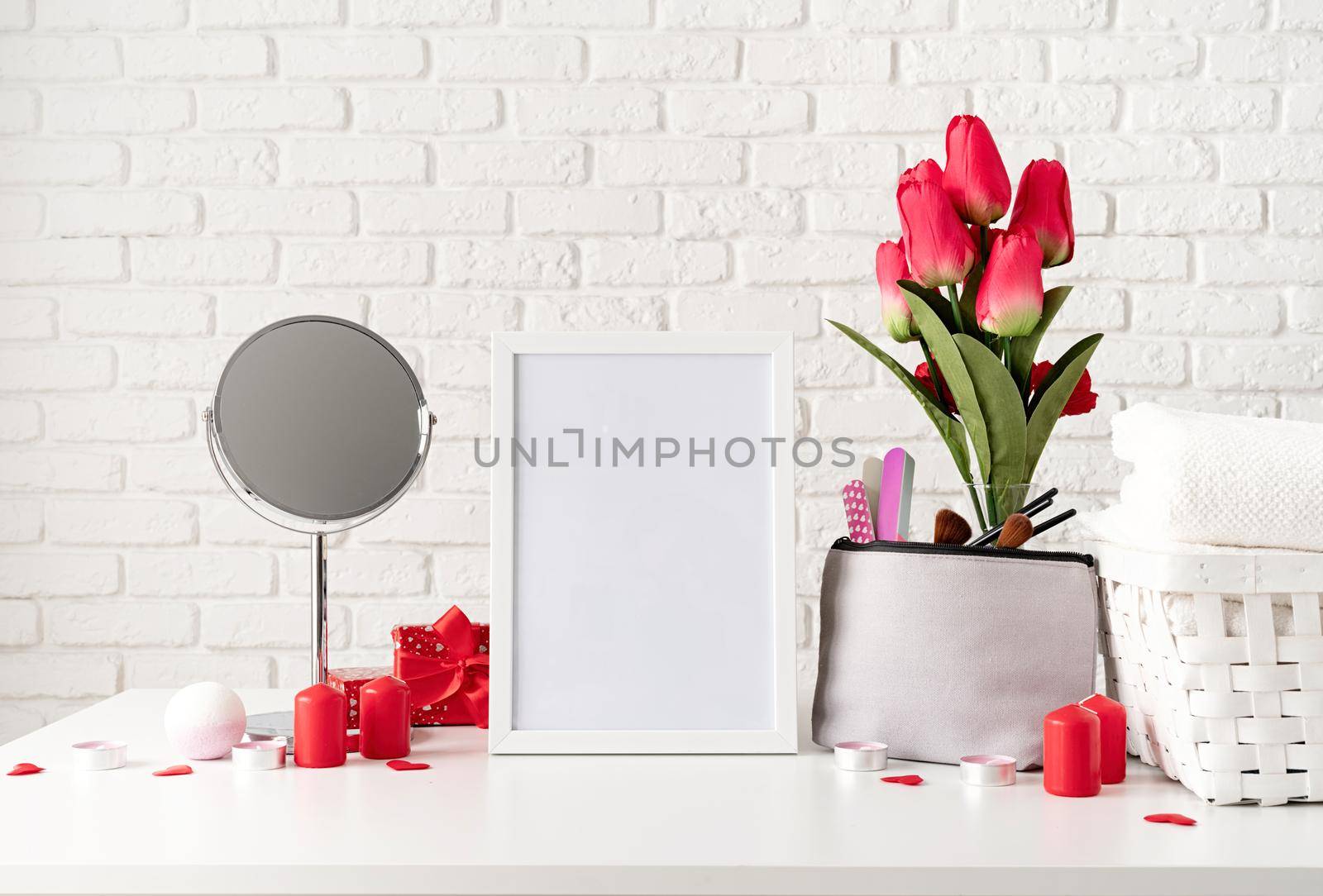 cosmetic and spa accessories with gift box, flowers and candles on dressing table front view on white brick wall background with blank frame for mock up by Desperada