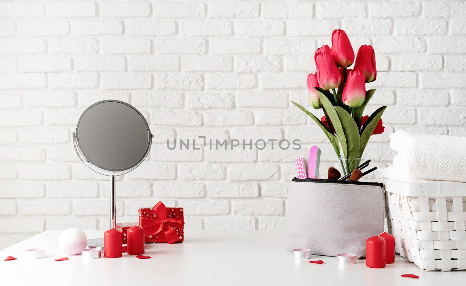 cosmetic and spa accessories with gift box, flowers and candles on dressing table front view on white brick wall background with copy space by Desperada