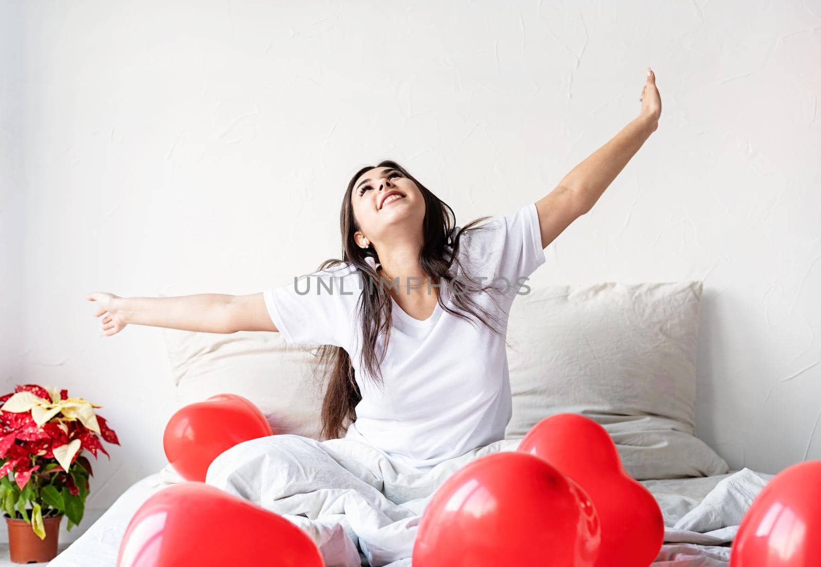 Young happy brunette woman sitting awake in the bed with red heart shaped balloons by Desperada
