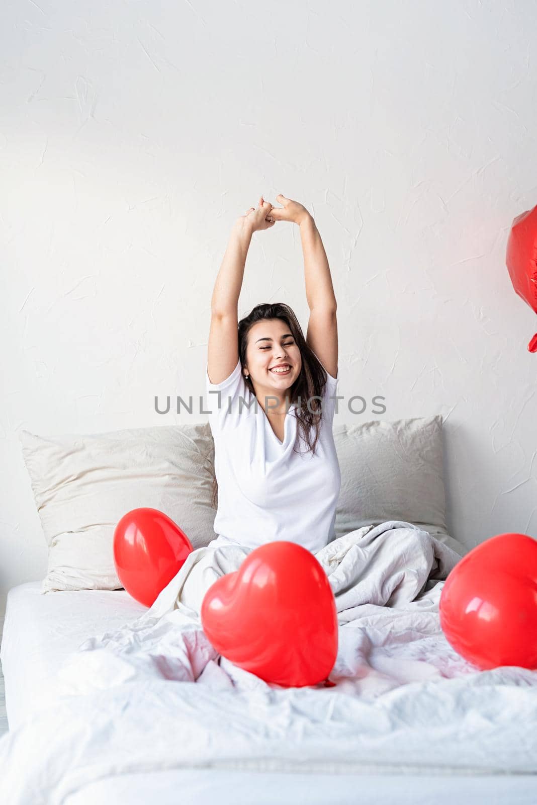 Young happy brunette woman sitting awake in the bed with red heart shaped balloons by Desperada