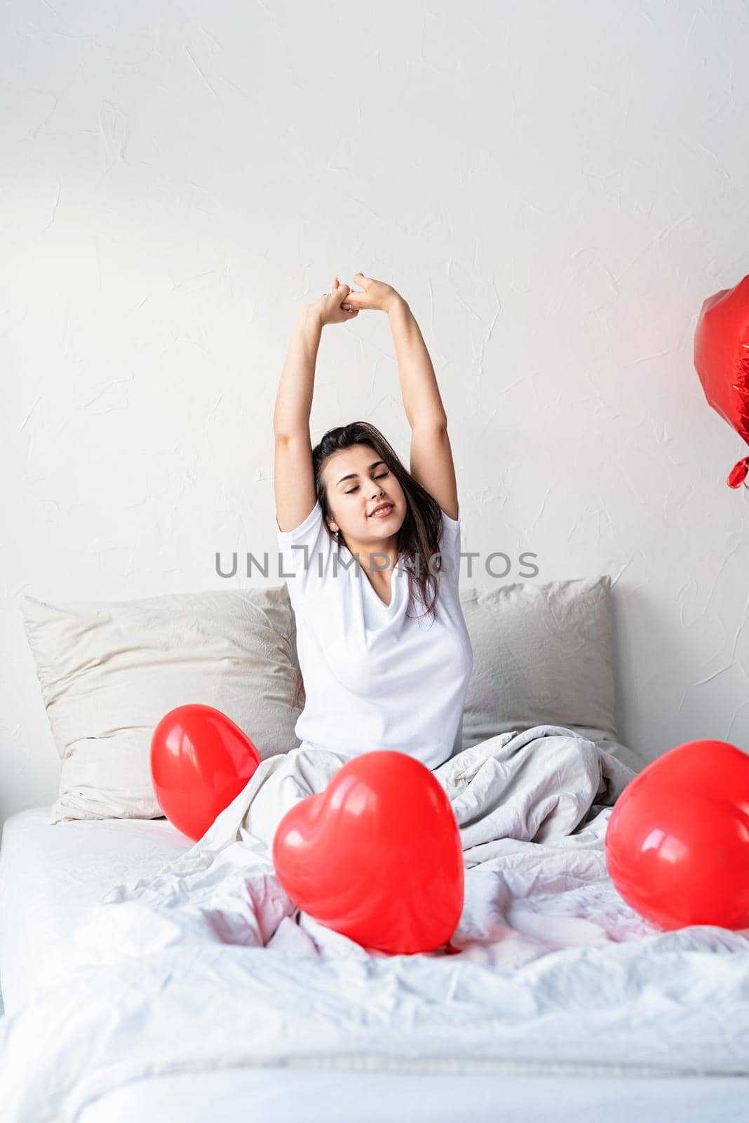 Valentine's Day. Sleeping. Young happy brunette woman sitting awake in the bed with red heart shaped balloons