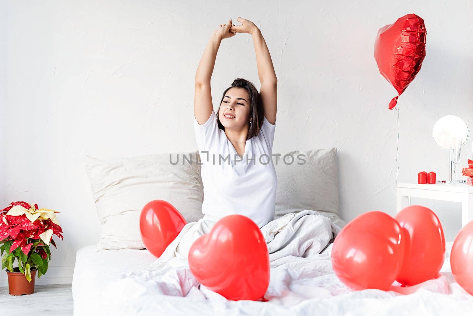 Valentine's Day. Sleeping. Young happy brunette woman sitting awake in the bed with red heart shaped balloons