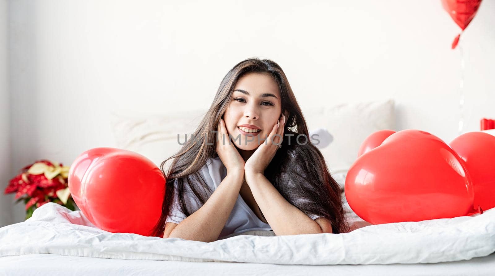 Young happy brunette woman laying in the bed with red heart shaped balloons by Desperada