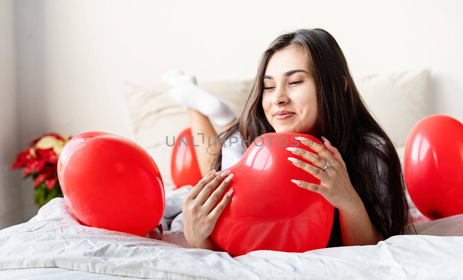 Young happy brunette woman laying in the bed with red heart shaped balloons by Desperada