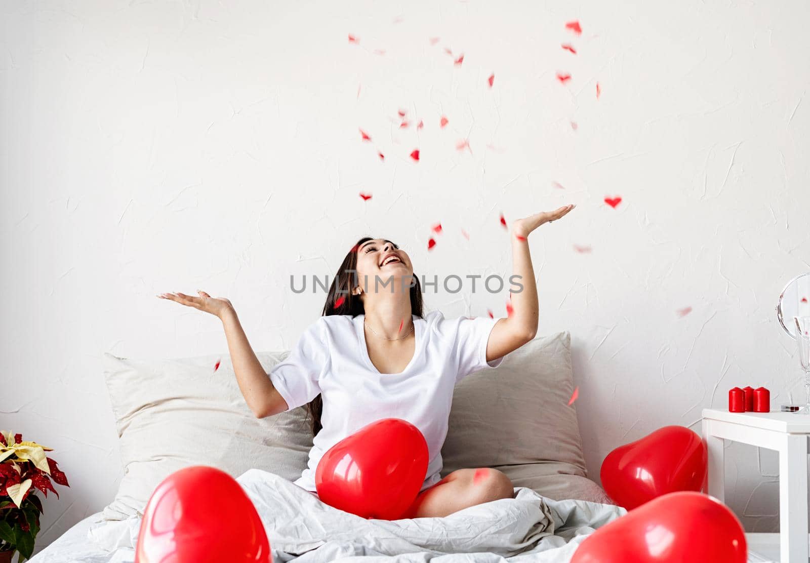 Valentine's Day. Sleeping. Young happy brunette woman sitting in the bed with red heart shaped balloons throwing confetti in the air
