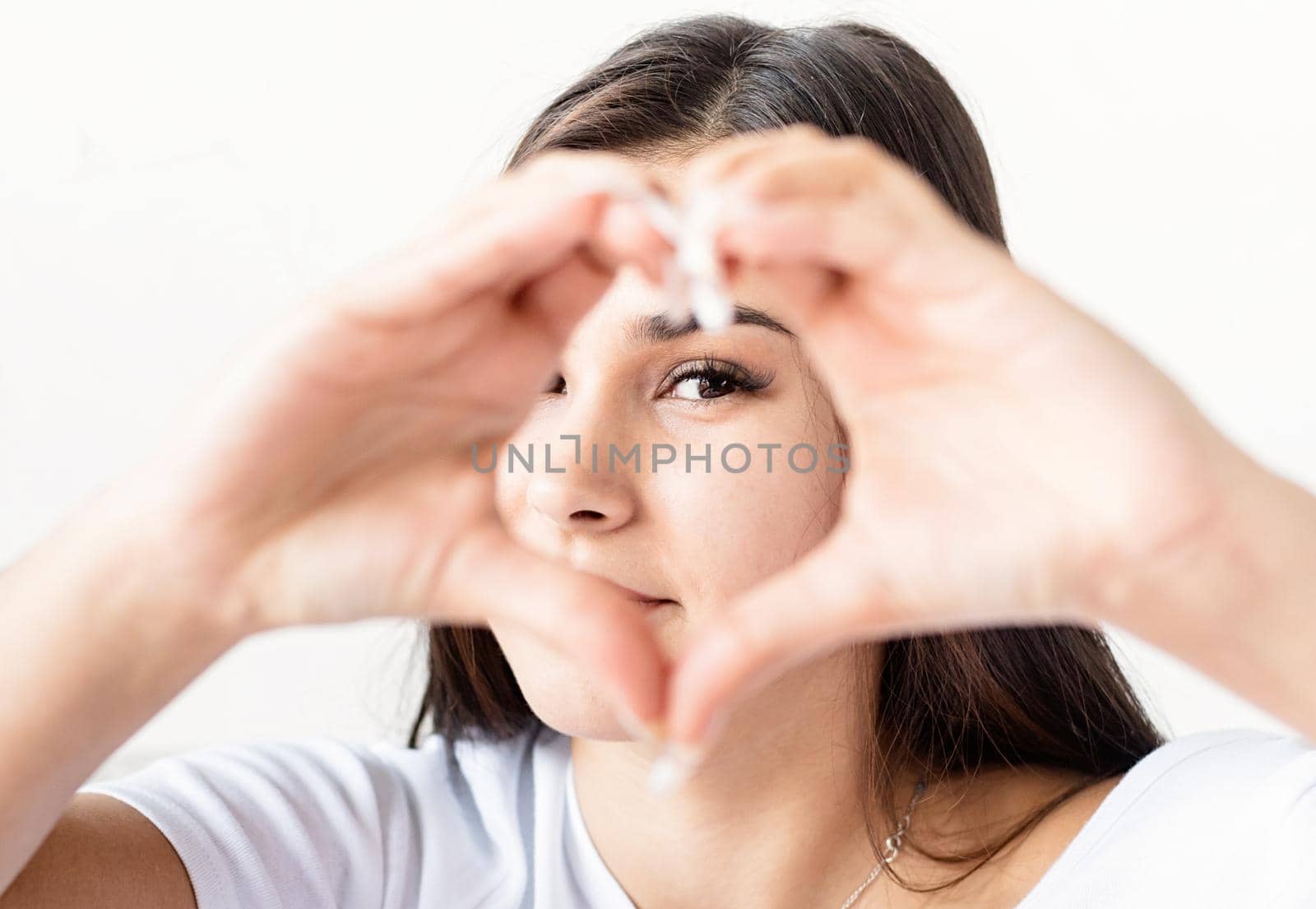 Young happy brunette woman in white t-shirt showing heart sign with her hands in front of face by Desperada