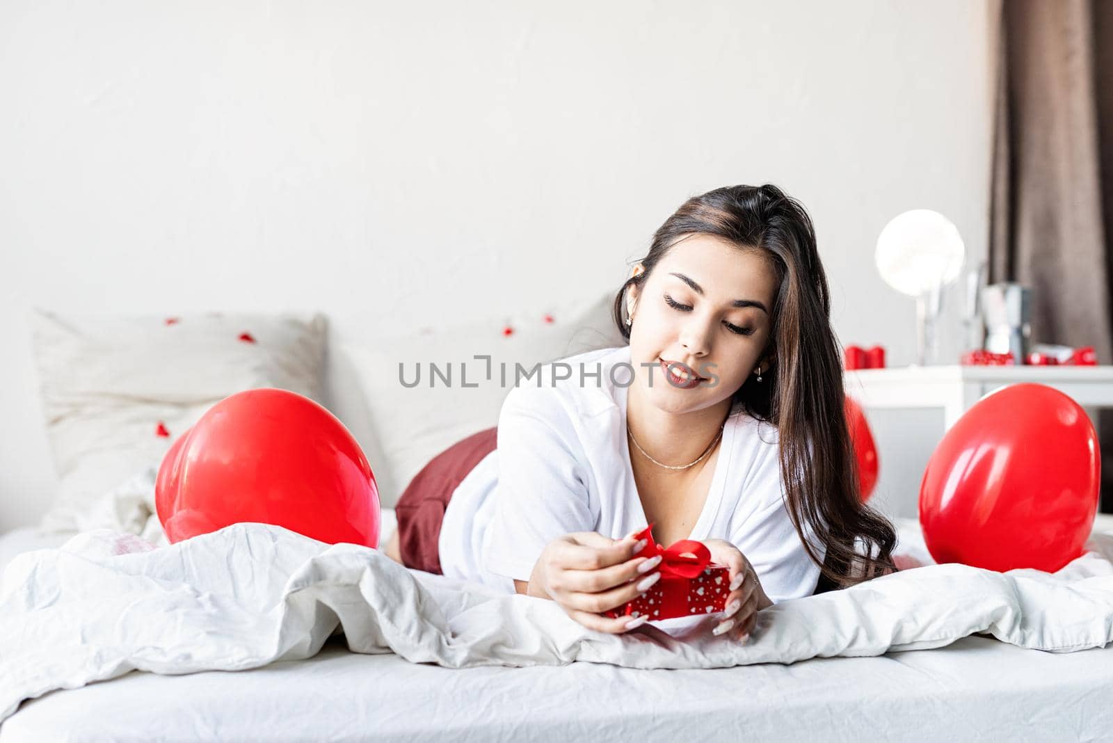 Young happy brunette woman laying in the bed with red heart shaped balloons unwrapping the present by Desperada