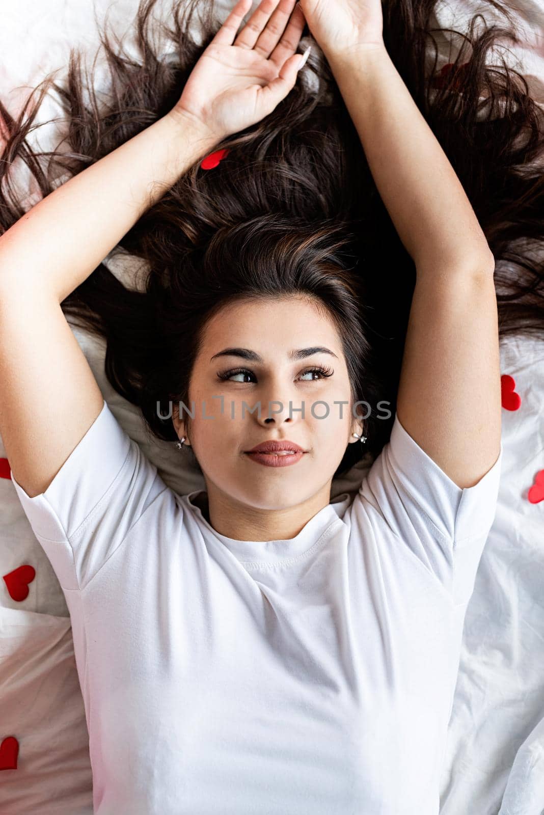 Valentines Day. Young happy brunette woman laying in the bed with red heart shaped confetti