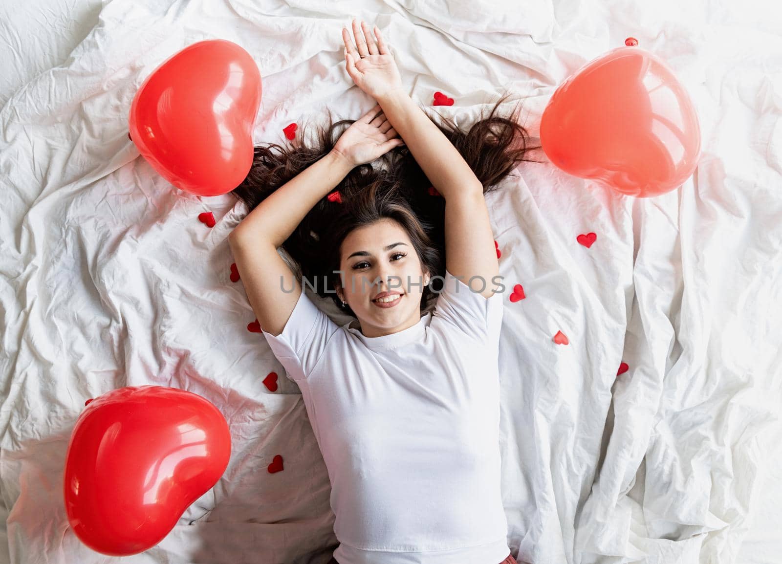Valentines Day. Young happy brunette woman laying in the bed with red heart shaped balloons and decorations