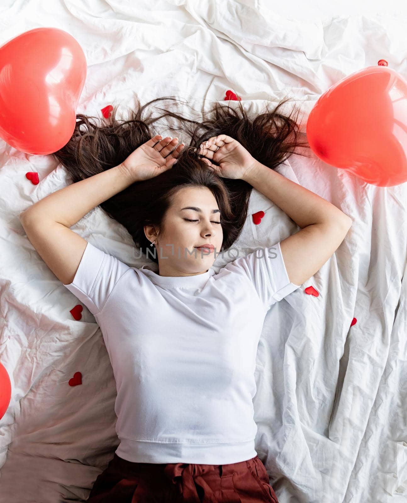 Valentines Day. Young happy brunette woman laying in the bed with red heart shaped balloons and decorations