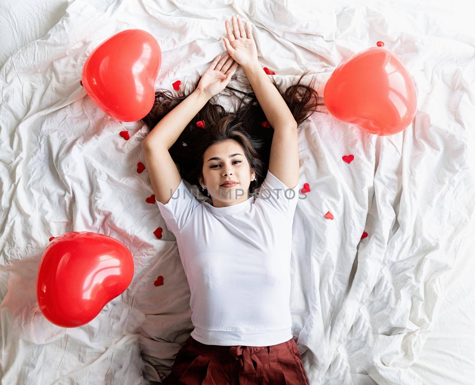 Valentines Day. Young happy brunette woman laying in the bed with red heart shaped balloons and decorations