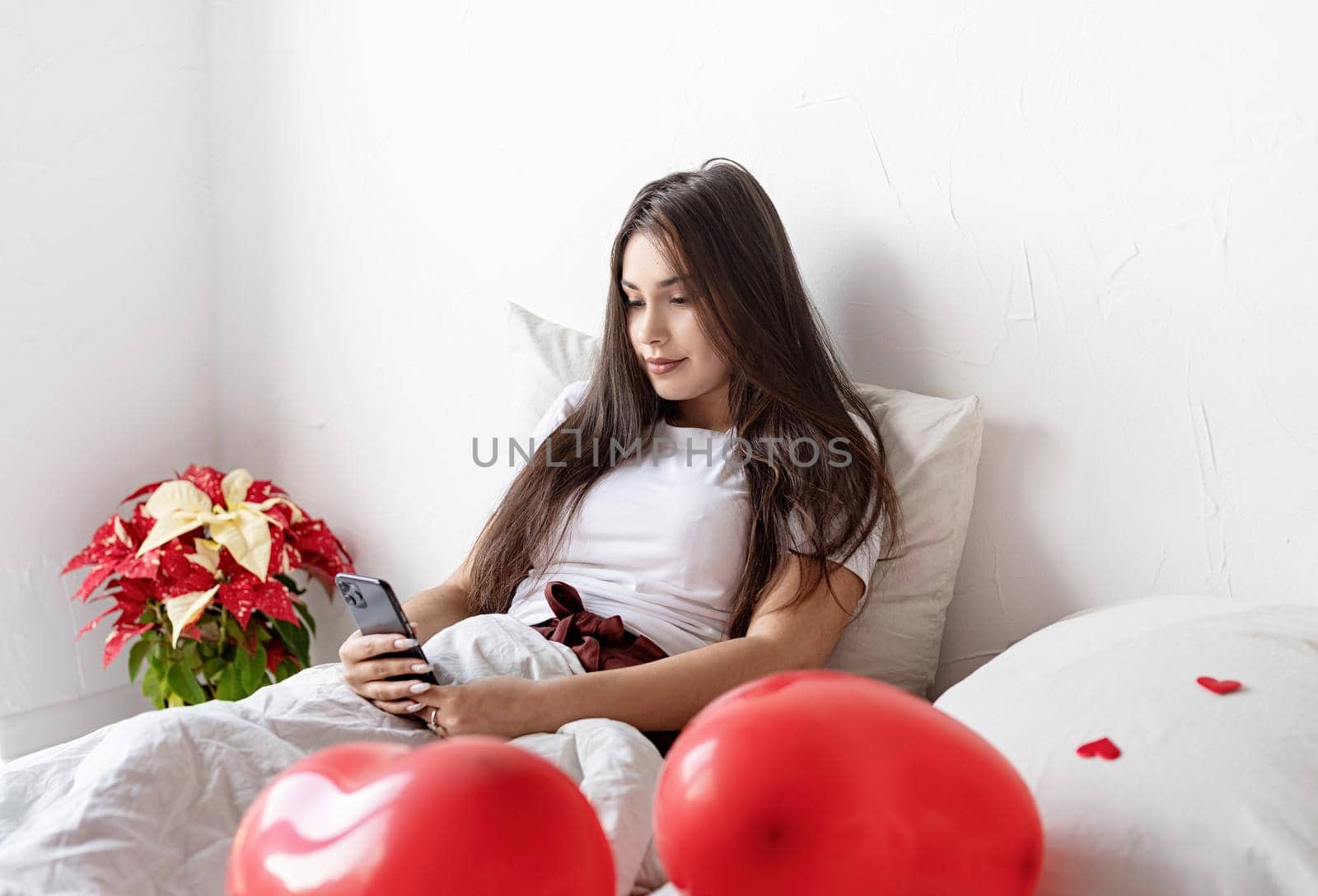 Valentines Day. Young brunette woman sitting awake in the bed with red heart shaped balloons and decorations texting