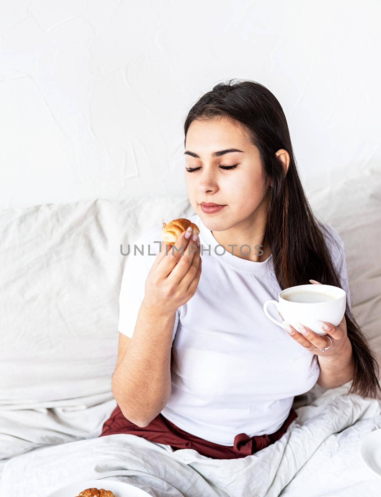 Young brunette woman sitting in the bed with eating croissants by Desperada