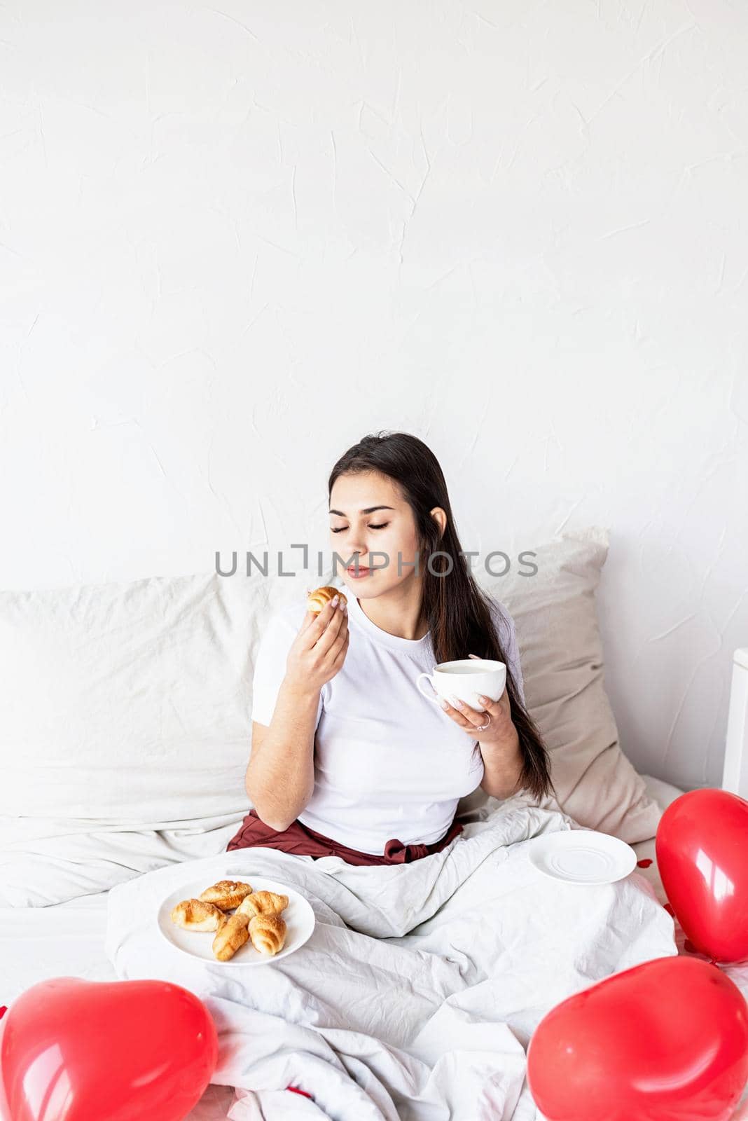 Young brunette woman sitting awake in the bed with red heart shaped balloons and decorations drinking coffee eating croissants by Desperada