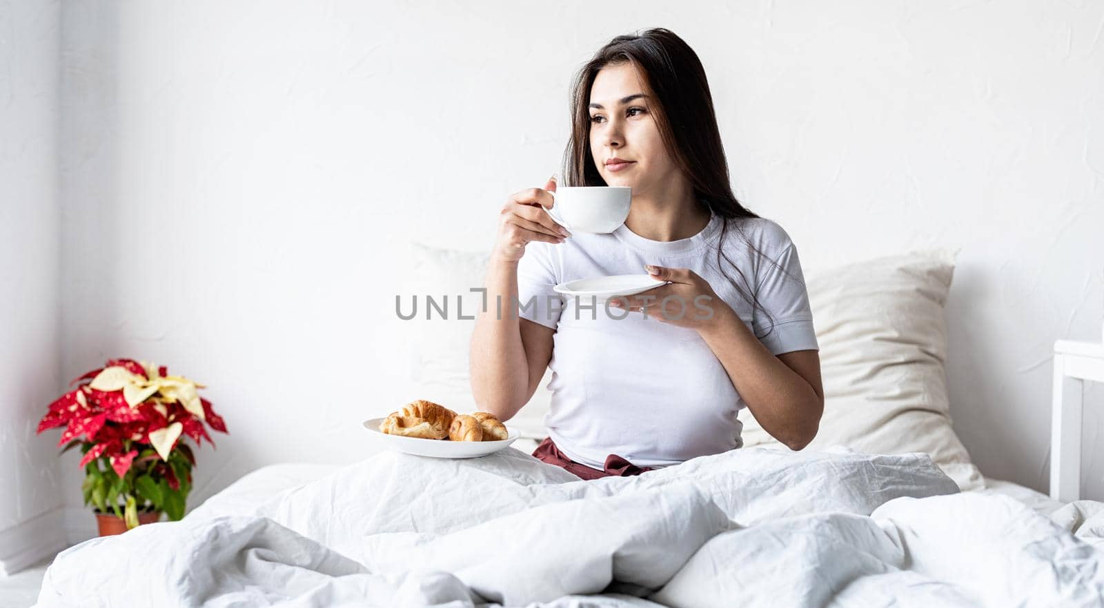 Valentines Day. Young brunette woman sitting awake in the bed with red heart shaped balloons and decorations drinking coffee eating croissants