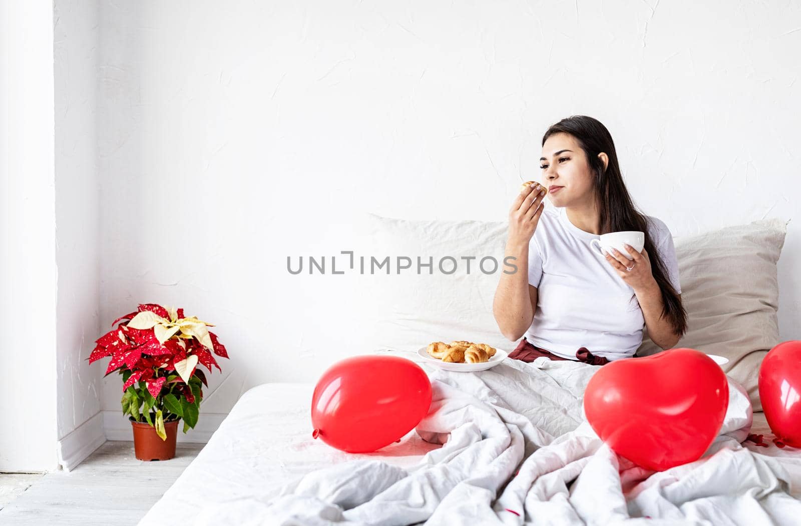 Valentines Day. Young brunette woman sitting awake in the bed with red heart shaped balloons and decorations drinking coffee eating croissants