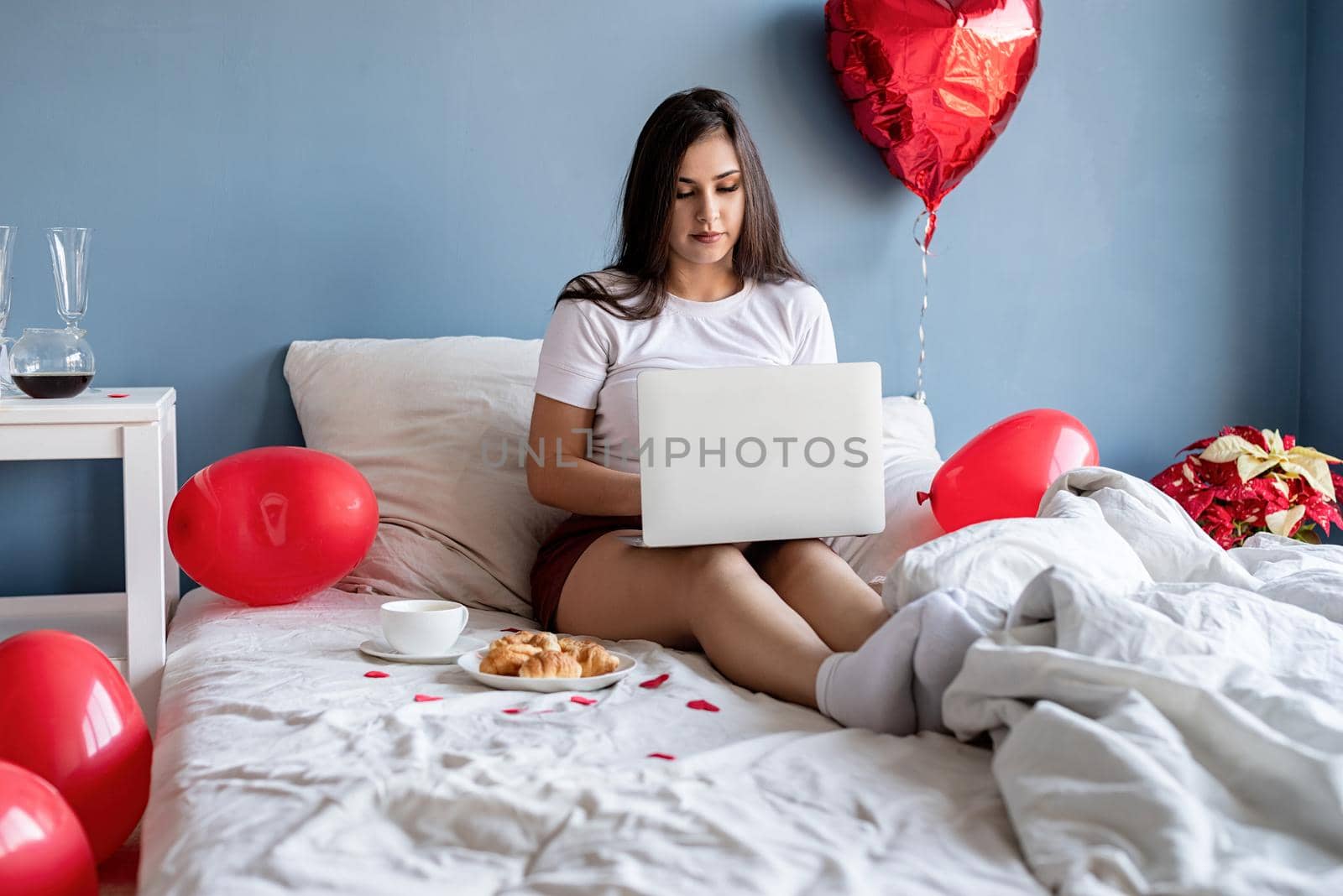 Young happy brunette woman sitting in the bed with red heart shaped balloons working on the laptop by Desperada