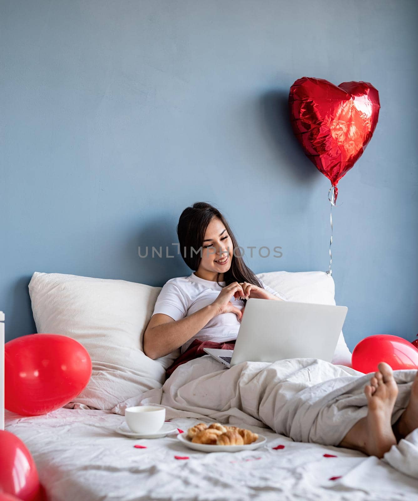 Valentines Day. Young happy brunette woman sitting in the bed with red heart shaped balloons chatting with her boyfriend on laptop showing heart gesture with hands