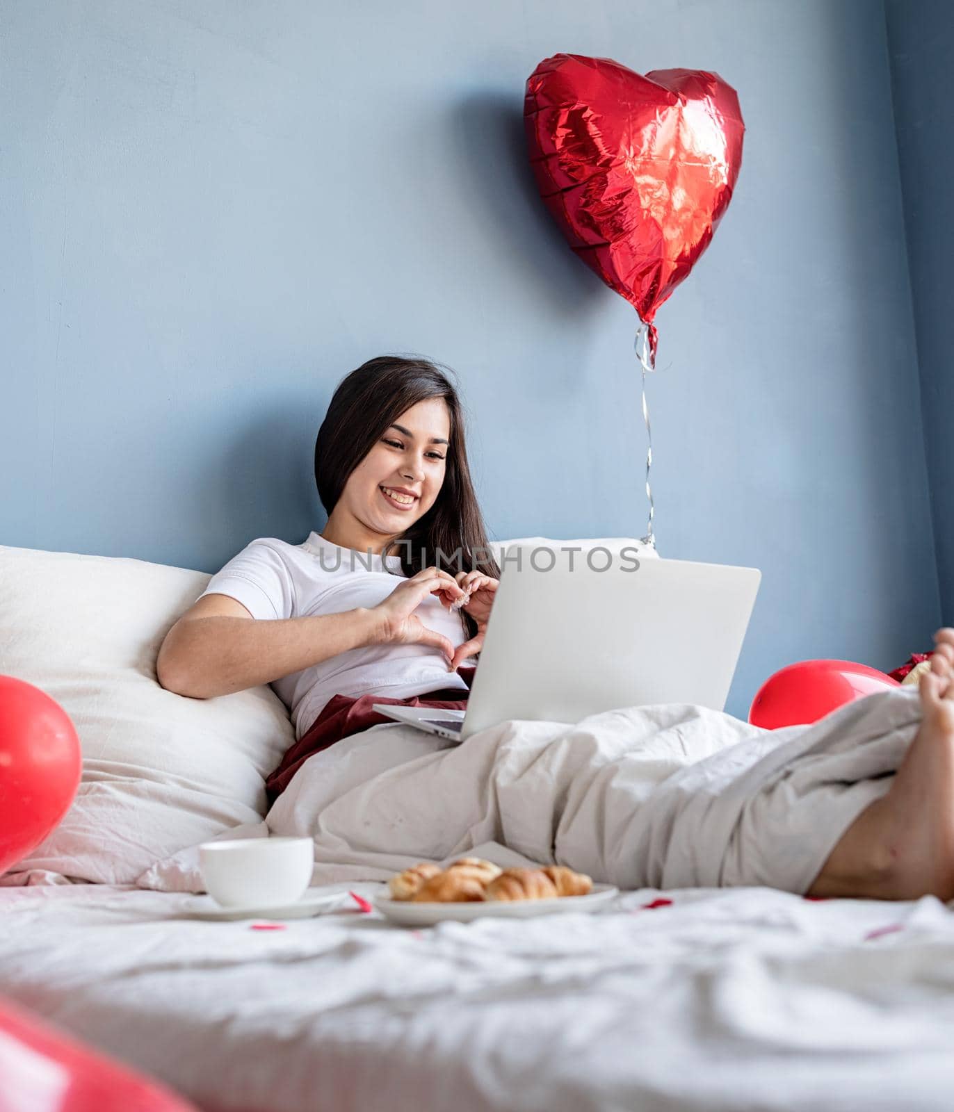 Valentines Day. Young happy brunette woman sitting in the bed with red heart shaped balloons chatting with her boyfriend on laptop showing heart gesture with hands