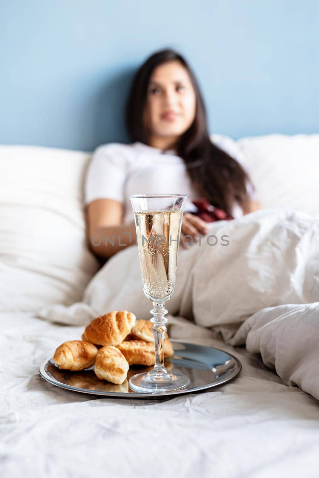 Valentines Day. Young brunette woman sitting awake in the bed with red heart shaped balloons and decorations drinking champagne eating croissants