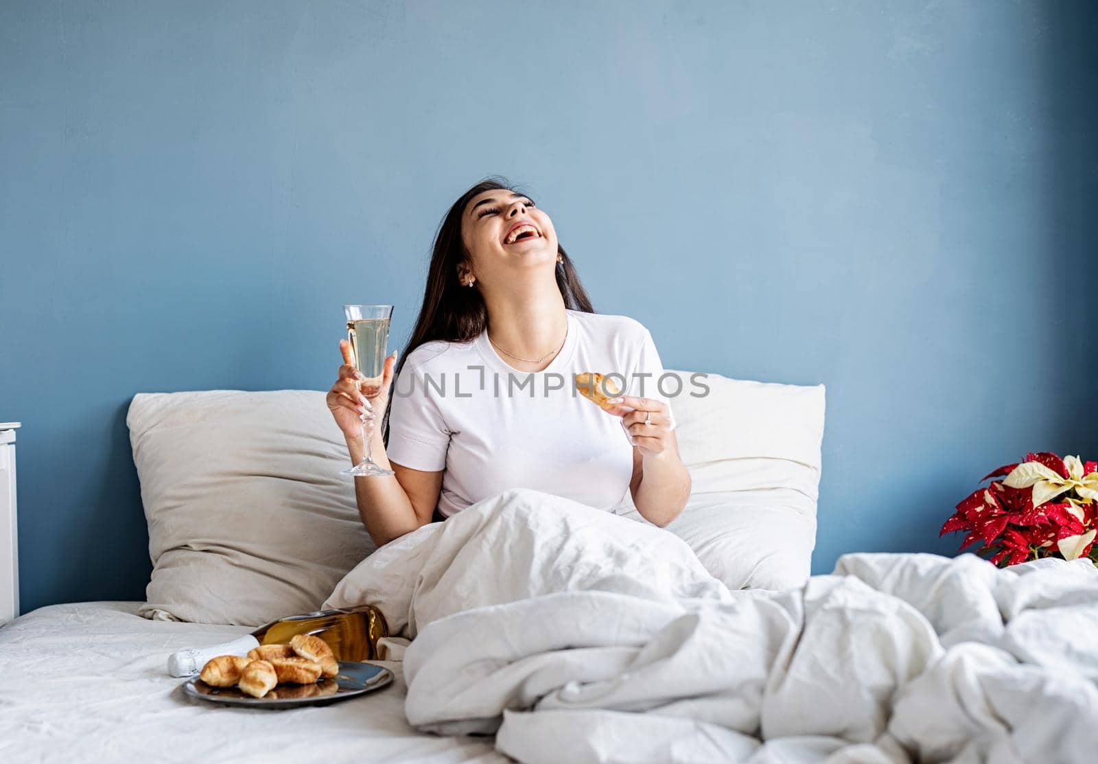 Young brunette woman sitting awake in the bed with red heart shaped balloons and decorations drinking champagne eating croissants by Desperada