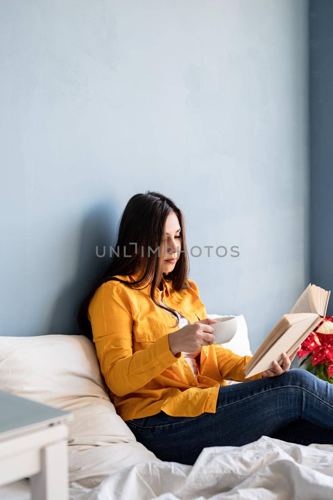 Young brunette woman sitting in the bed with eating croissants and reading a book by Desperada
