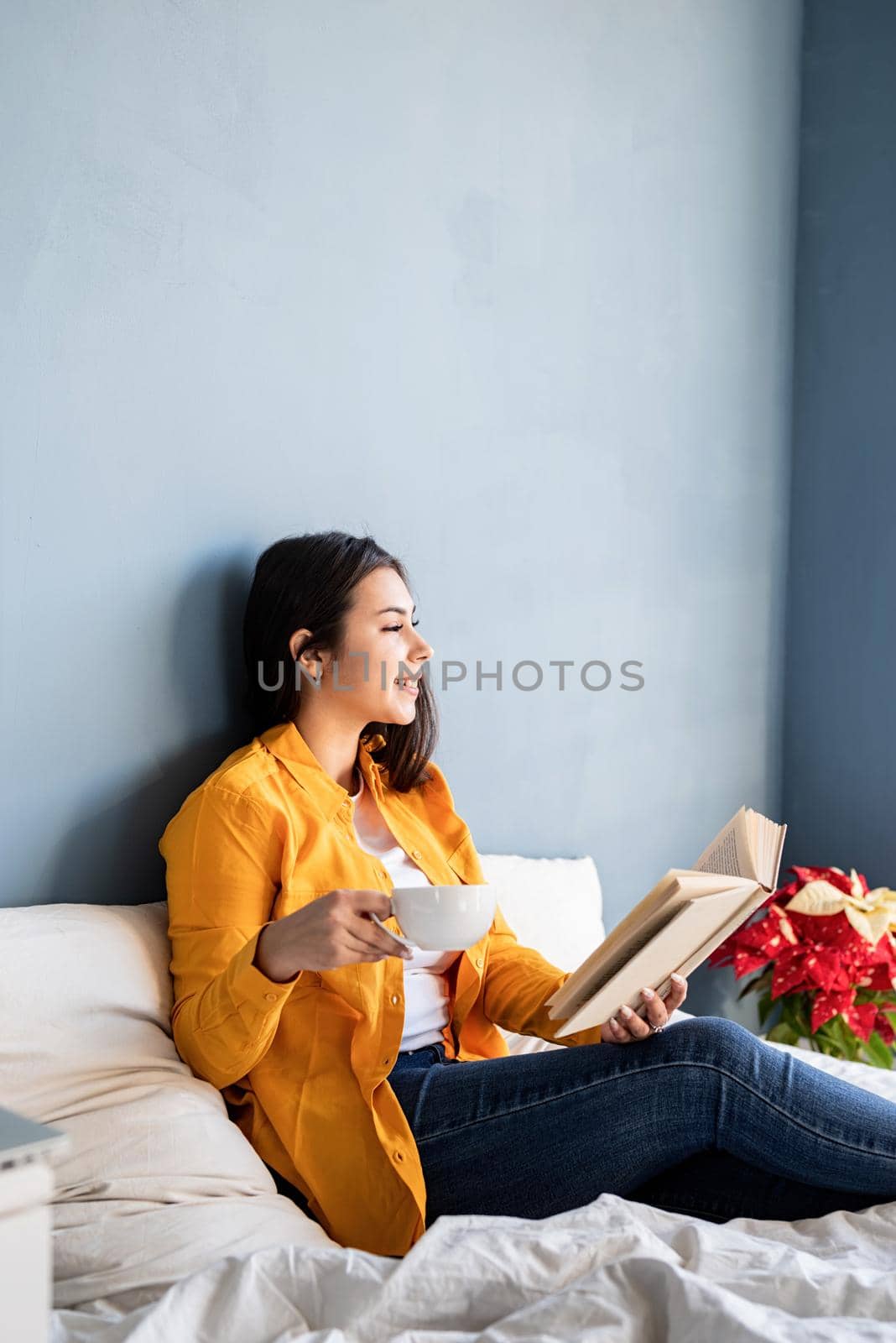 Young brunette woman sitting in the bed with eating croissants and reading a book by Desperada